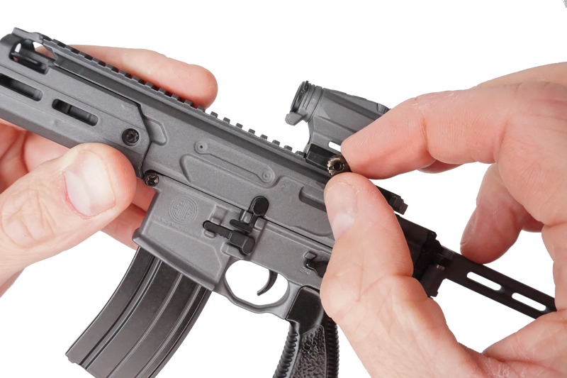 Close up of the stone grey SIG MCX model being held in two hands. The  left hand is holding the model and the right hand is adjusting the scope