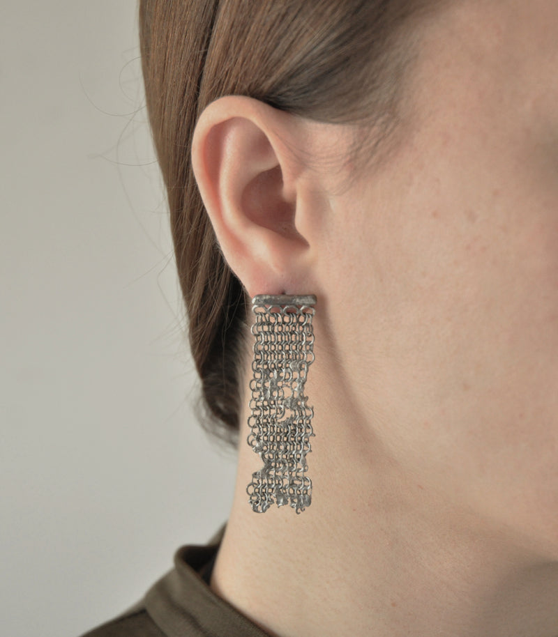 A model wearing a distressed silver chain mail drop earring with the appearance of being melted