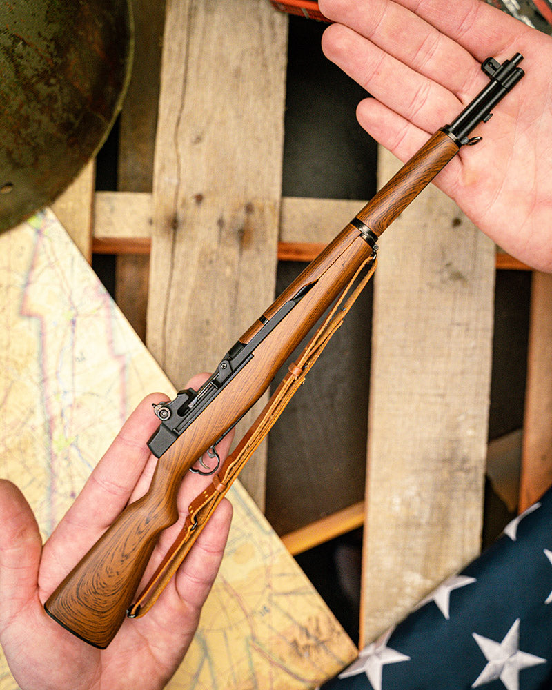M1 Garand Model being held in two hands, resting on its side, showing its scale. In the background a wooden pallet with a world war 2 helmet, a map, and an american flag can be seen.