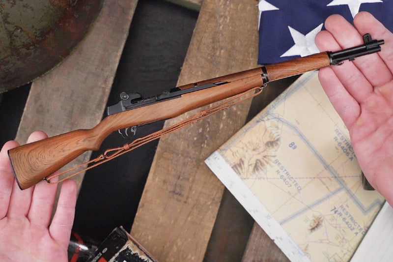 M1 Garand model being held on its side by two hands, indicating its small scale. On the workbench in the background is a world war 2 helmet, an american flag, and a map.