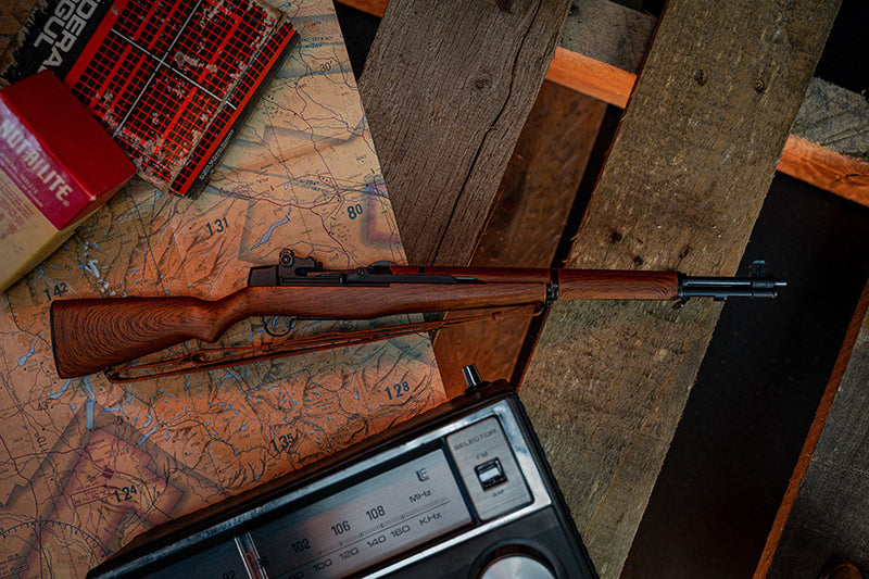 M1 Garand model resting on its side on a wooden pallet, facing towards the right. In the background there is also a map and a vintage radio