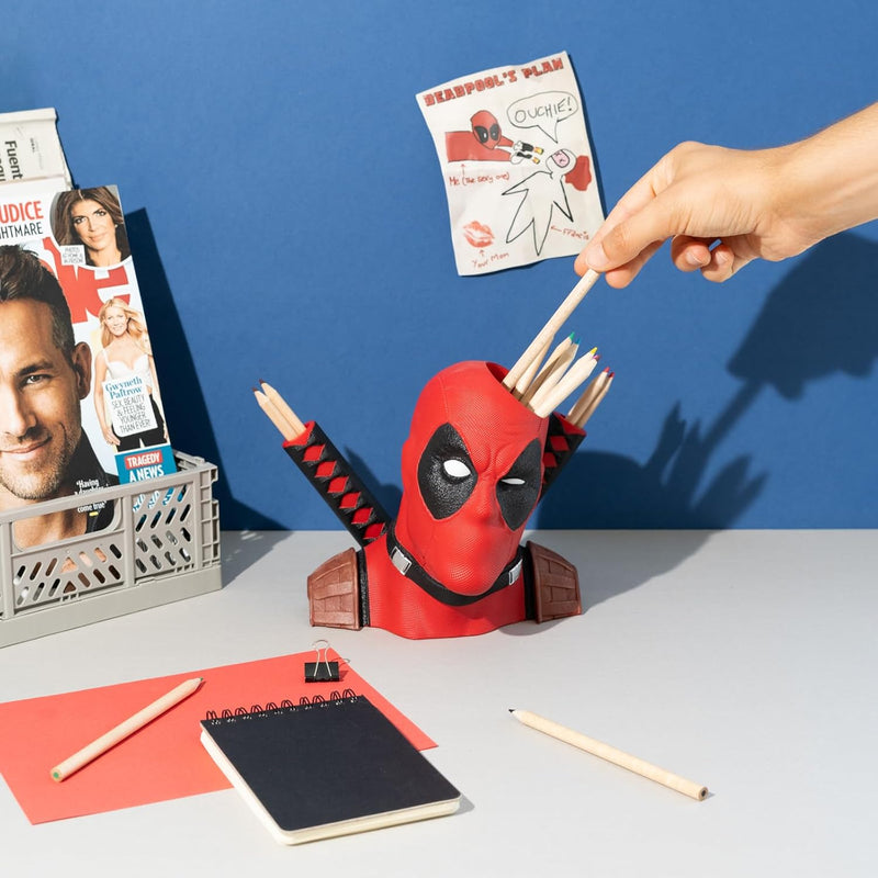 Deadpool-themed pencil holder with pencils on a desk against a blue wall.